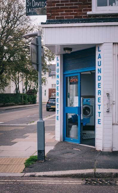 Exterior view of a small laundry facility with white wooden panel walls and bright blue signage reading 'LAUNDERETTE' on both sides of the open door. Inside, a commercial washing machine with a silver
