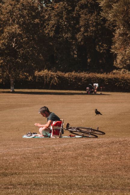 A woman with long red hair wearing a white and navy striped long-sleeve shirt and black pants is sitting cross-legged on a white blanket spread over a grassy area within Clissold Park. She is in the p