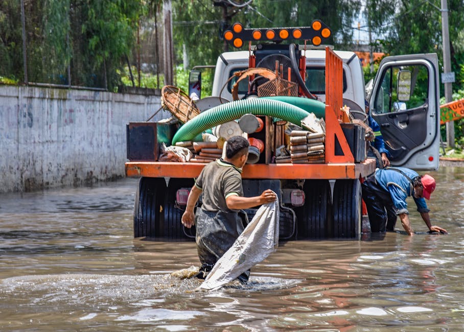 Photograph of a flood in an urban street showing a partially submerged black luxury sedan with water reaching halfway up its doors, and a white taxi with orange and black markings submerged up to its