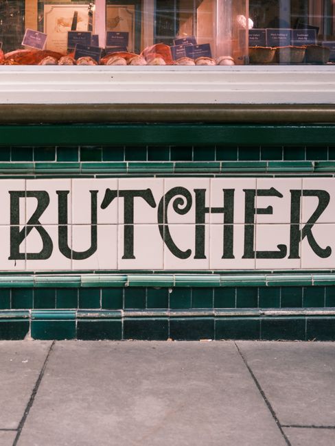 Close-up view of a tiled storefront with large black letters spelling 'BUTCHER' on white tiles framed by dark green tiles. Above the tiled signage, a glass display case shows raw meat cuts on butcher 